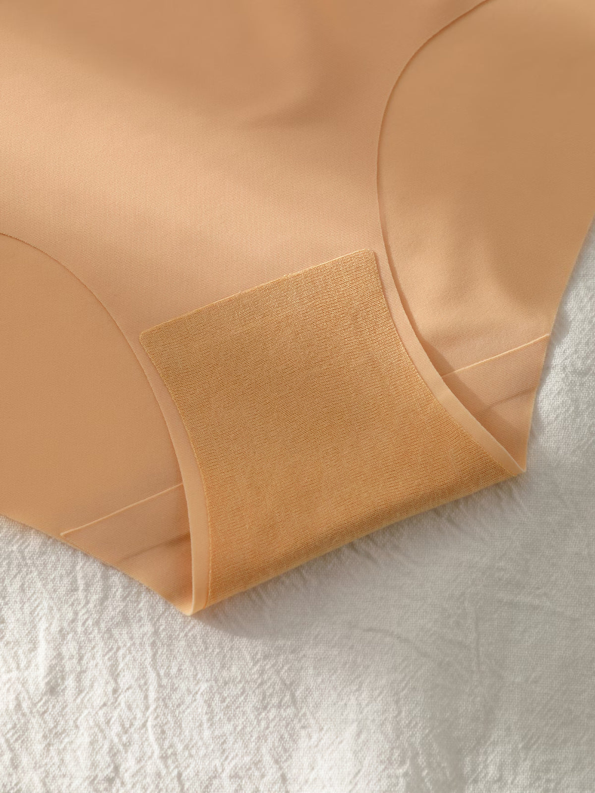 Close-up of a nude beige shapewear brief or underwear leg piece laid on a white textured fabric background, showing the fabric texture and curved leg contours.