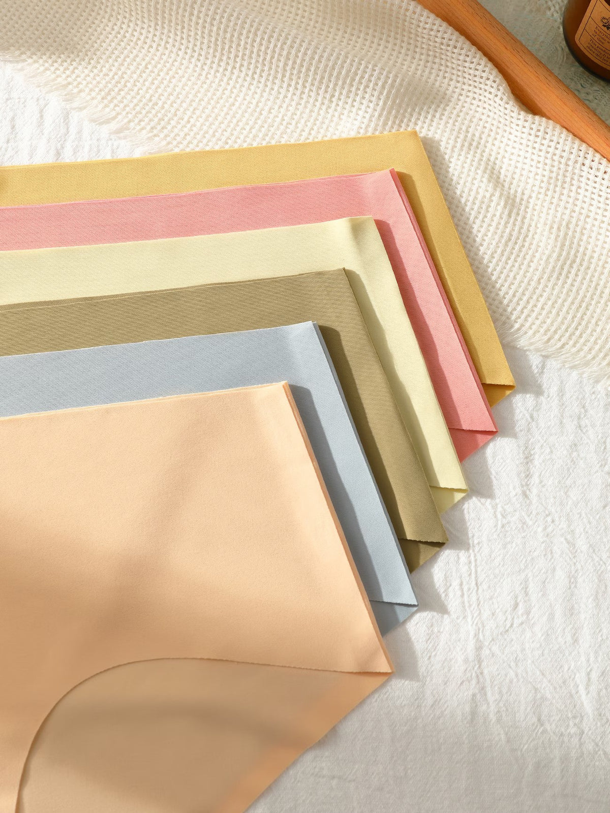 A fan arrangement of soft, solid-colored underwear or fabric pieces in peach, light blue, taupe, cream, pink, and mustard yellow, displayed on a textured white fabric background.