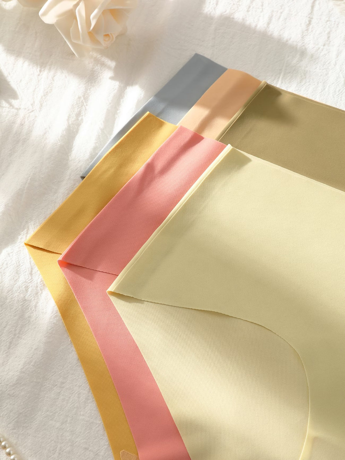 Four soft fabric panty in pastel shades of cream, pink, mustard yellow, and light blue, arranged diagonally on a white textured surface with soft shadows.