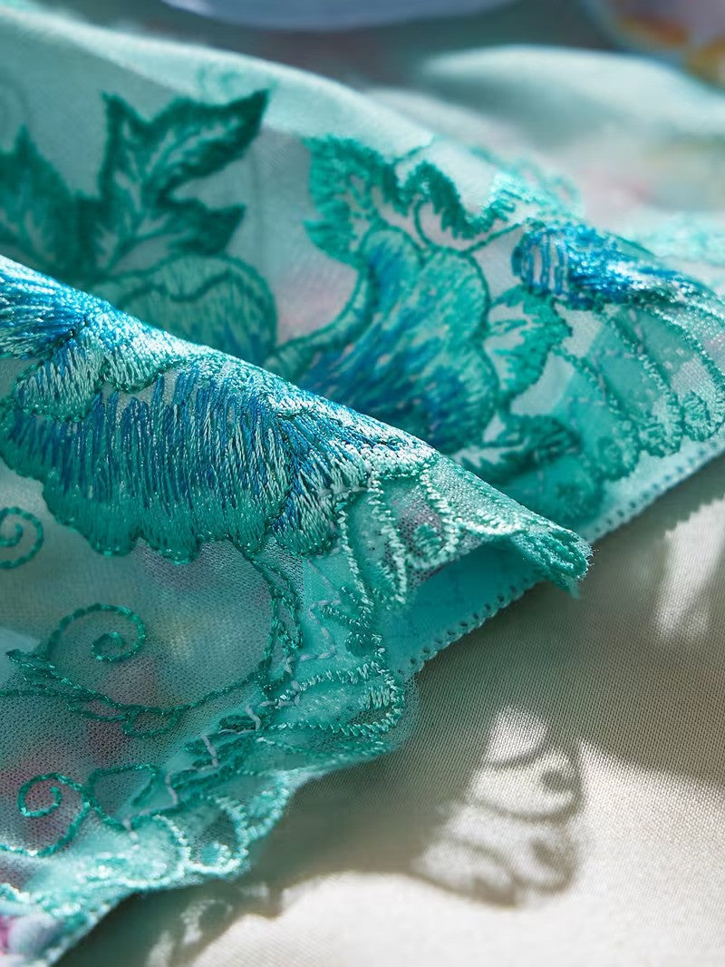 A close-up of sheer lace fabric with teal and turquoise floral embroidery and metallic thread detailing, draped over a light neutral surface.