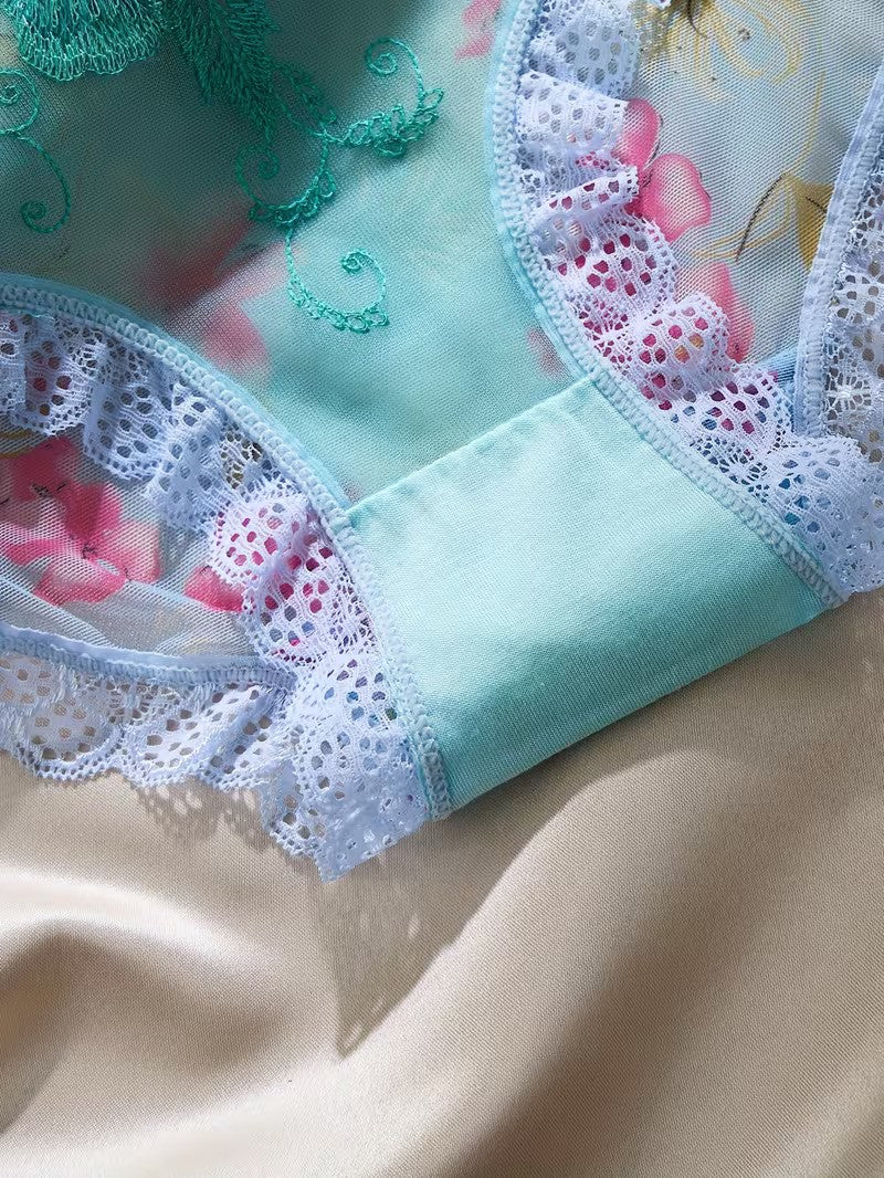 A close-up of a light blue lace-trimmed garment laid on a beige satin fabric. The fabric has a floral print with pink flowers and green embroidered swirls.