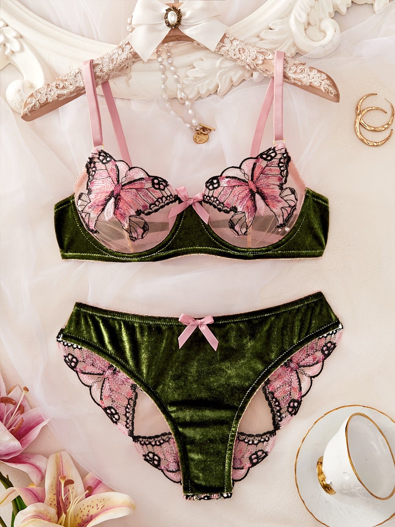A two-piece lingerie set featuring a velvet green bra and matching panties with pink and black butterfly embroidery and lace trim, displayed on a hanger and fabric backdrop with decorative props.