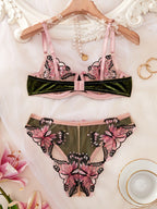 A two-piece lingerie set featuring a pink and dark green velvet bralette with butterfly applique, and matching panties, displayed on a wooden hanger with decorative props.