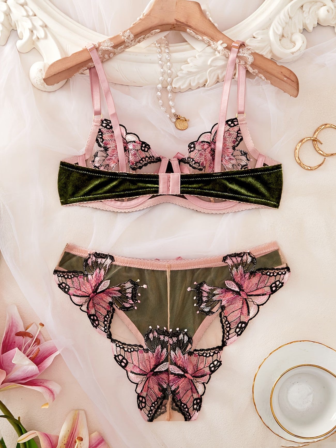 A two-piece lingerie set featuring a pink and dark green velvet bralette with butterfly applique, and matching panties, displayed on a wooden hanger with decorative props.