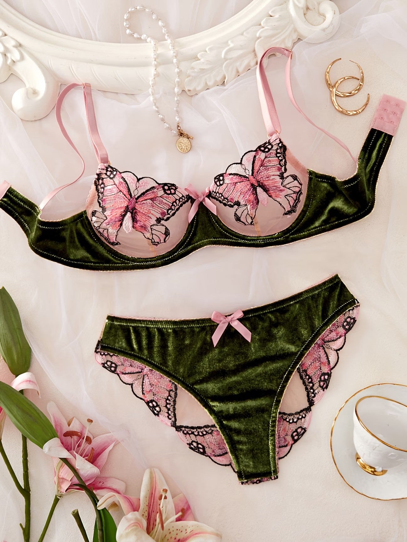 A two-piece lingerie set laid flat on a white fabric background, featuring an olive green velvet bra and matching panties with pink and black butterfly and lace details. The set is styled with a pearl necklace, gold rings, and pink lilies.
