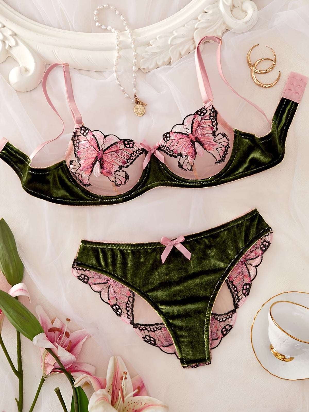 A two-piece lingerie set laid flat on a white fabric background, featuring an olive green velvet bra and matching panties with pink and black butterfly and lace details. The set is styled with a pearl necklace, gold rings, and pink lilies.