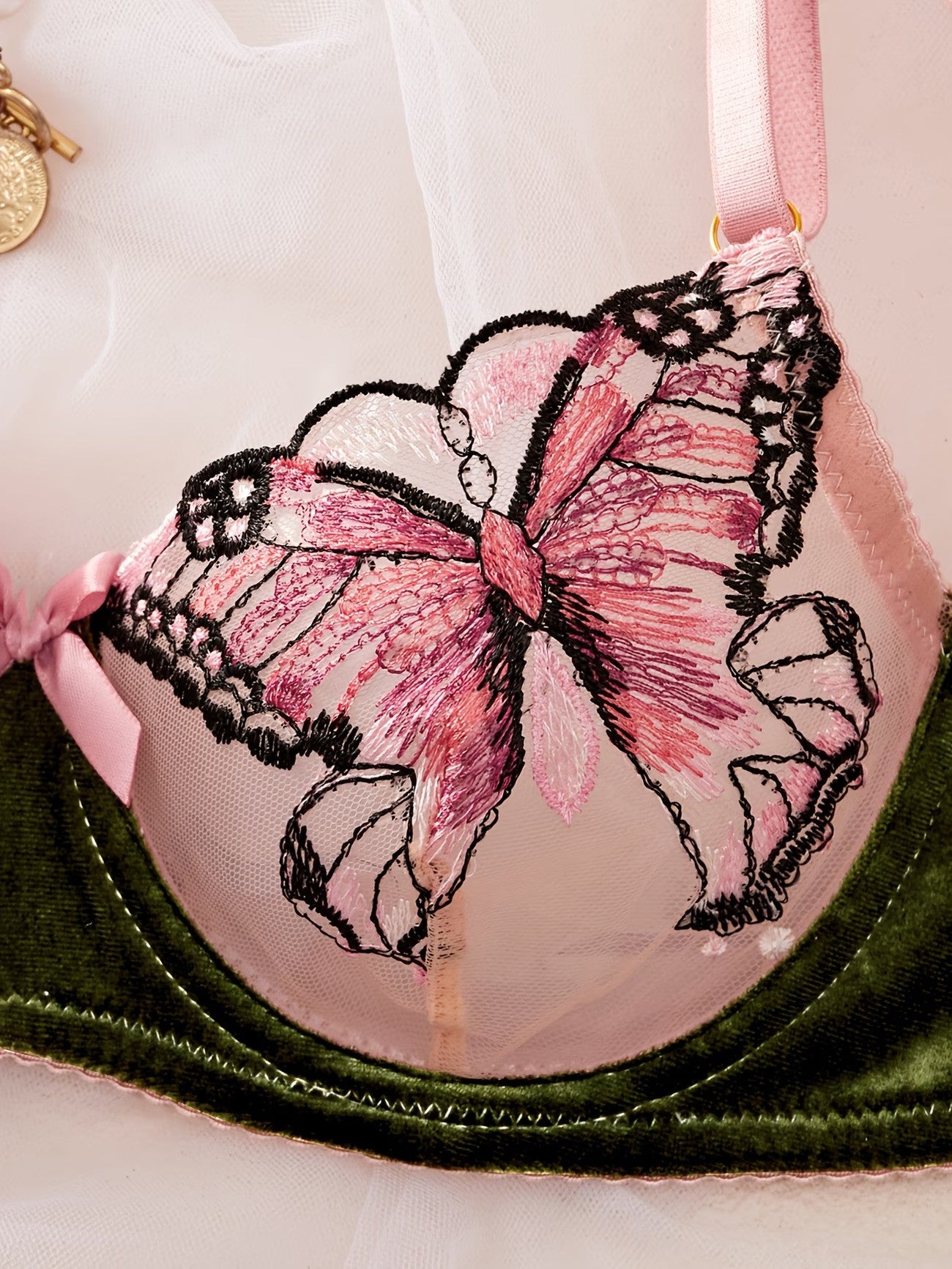 Close-up of a pink and green underwire bra featuring a large embroidered pink bow with black lace trim on the cup, with a pink ribbon strap and a dark green velvet band.