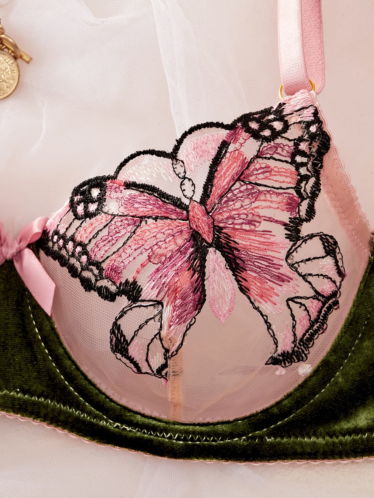 Close-up of a pink and green underwire bra featuring a large embroidered pink bow with black lace trim on the cup, with a pink ribbon strap and a dark green velvet band.